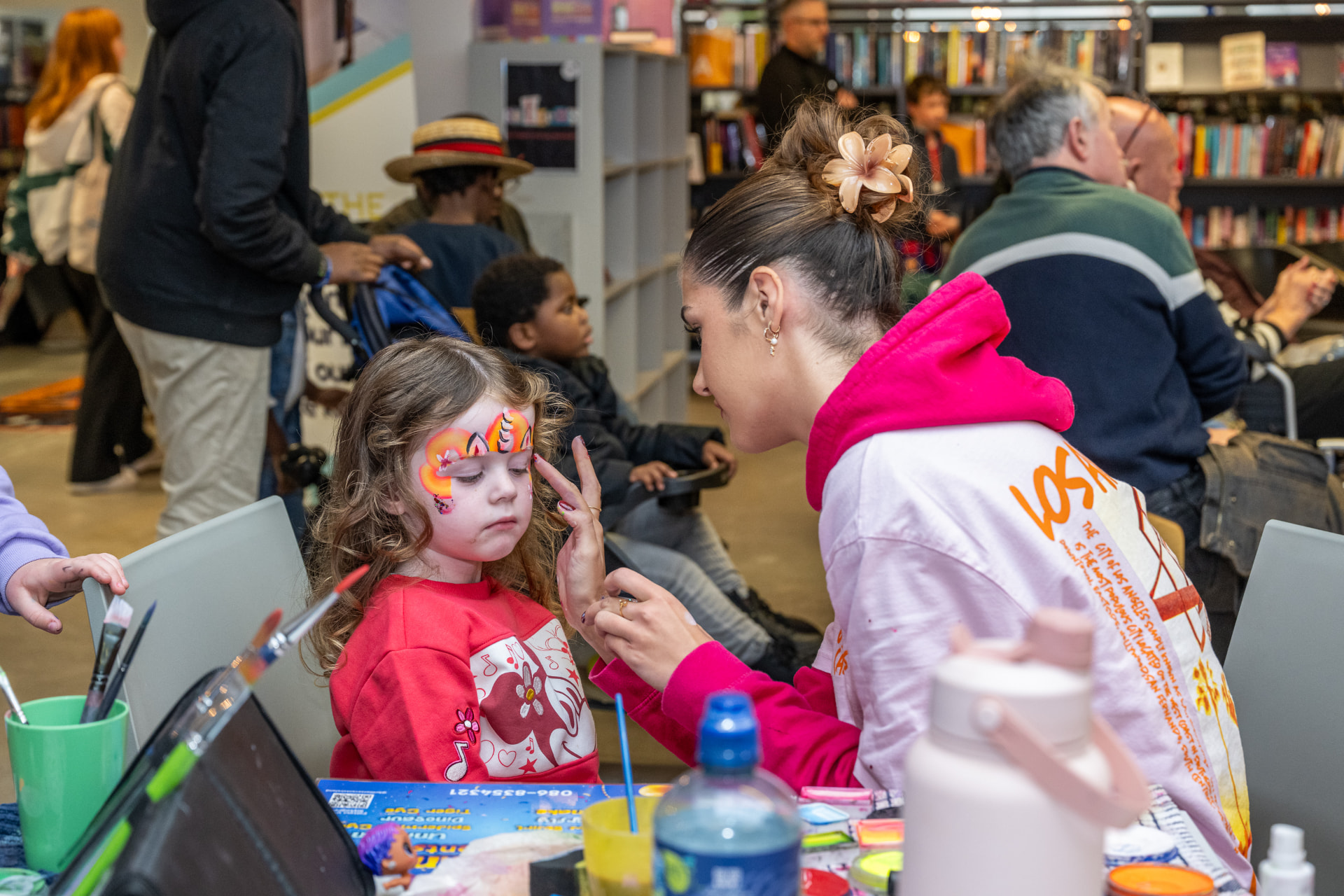 A face painter at a Red Line Book Festival Family Fun Day carefully painting a tiger design on a smiling child in a library surrounded by bookshelves.